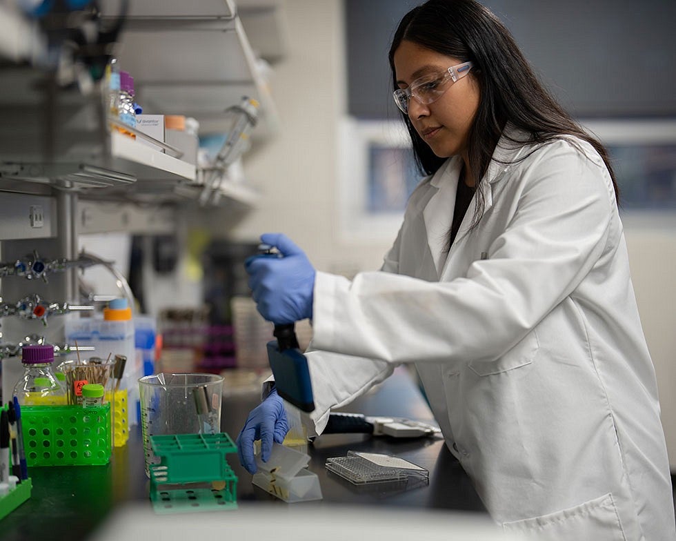 Researcher wearing white lab coat, gloves, and protective goggles working in lab