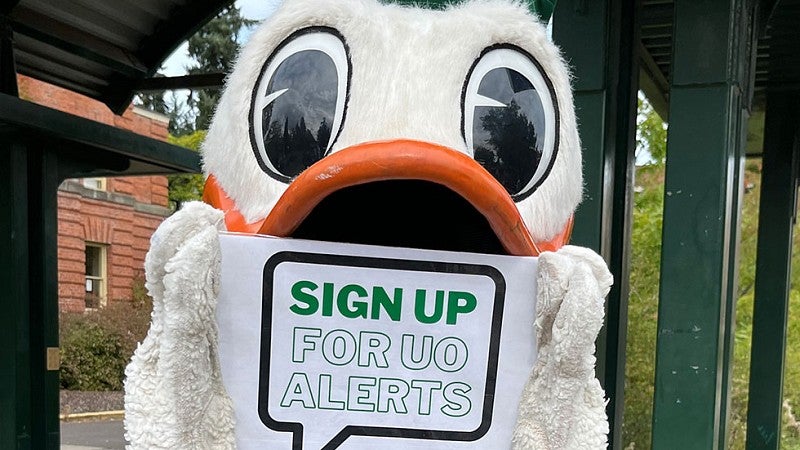 The Duck holding a sign reminding people to sign up for UO Alerts