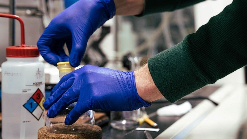 Close up of gloved hands unstopping a container in a lab, background has another container with hazard label