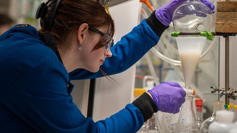 Researcher in chemistry lab wearing blue lab coat, gloves, eye protection while working