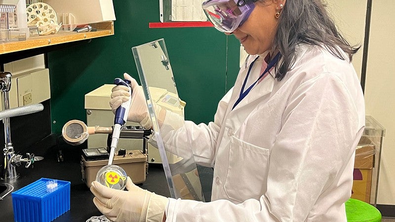 Researcher wearing eye protection, gloves, lab coat working behind radiation shield. Using geiger counter, pipettes, and other lab equipment. 